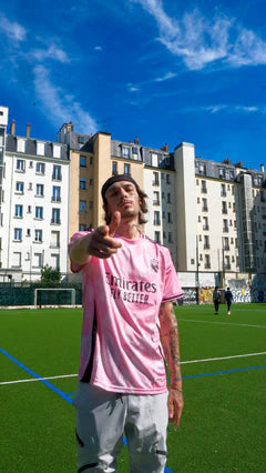 Jeune homme portant un maillot de foot rose sur un terrain urbain, immeubles en fond, ciel bleu.